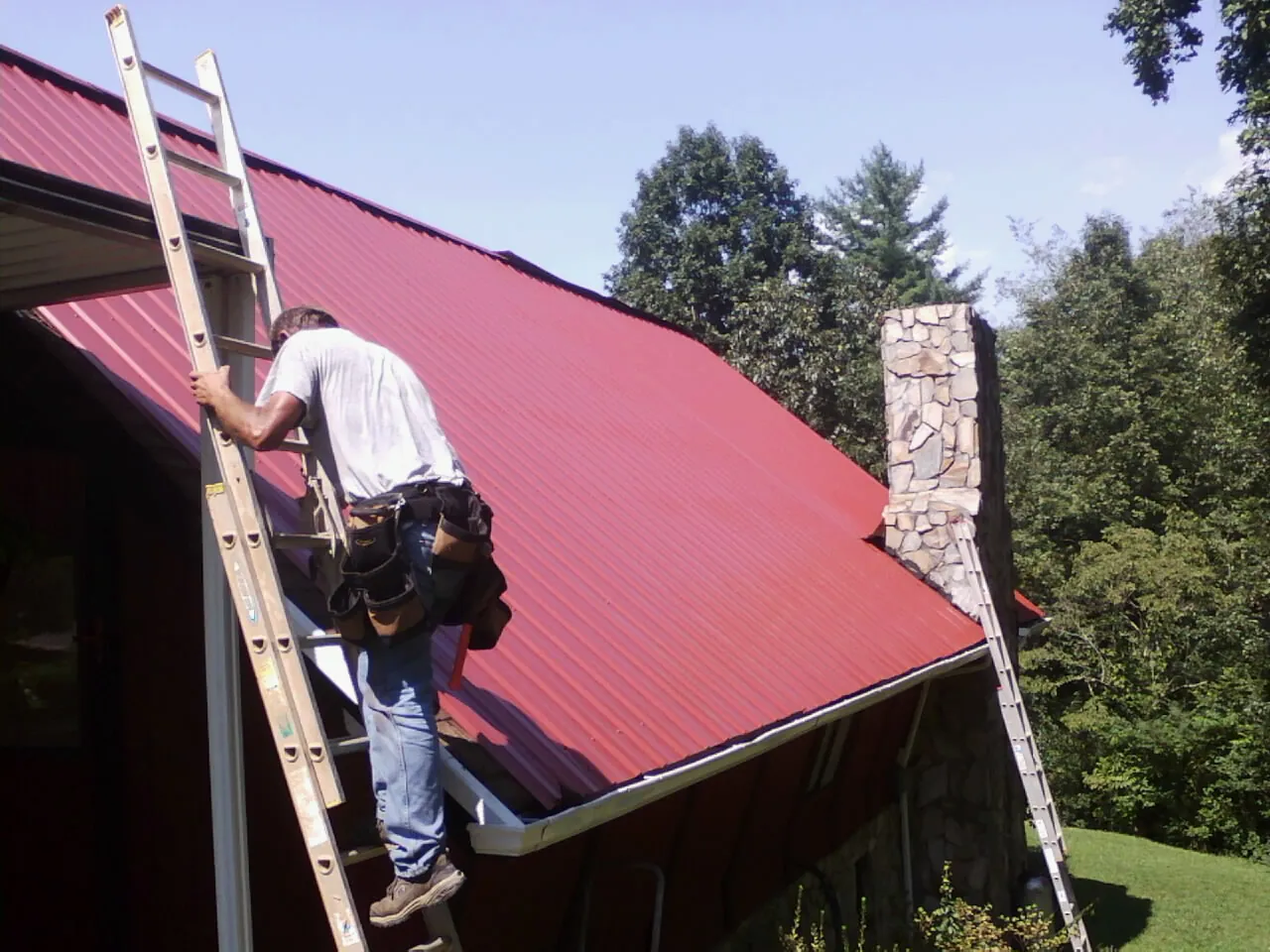 Roofer on a ladder beside a red metal roof and stone chimney — Soffit & Fascia Repair by Roofing Brothers in Avilla